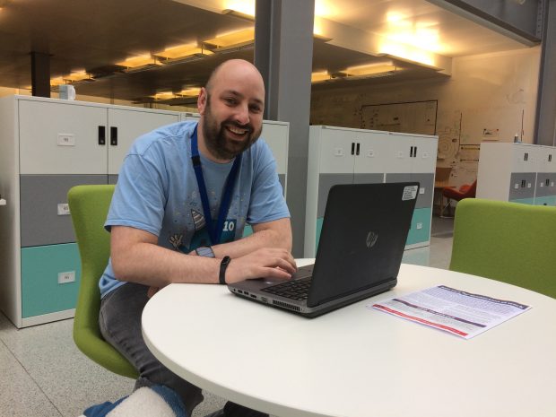 Adam Pearce sitting at a table in the Newcastle hub atrium smiling at the camera with one hand on his laptop