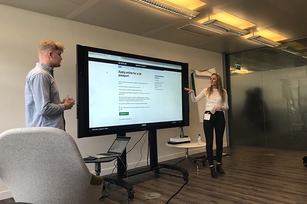 Two students standing at either side of a large display screen. On the screen is displayed the homepage for the Apply online for a UK passport website f