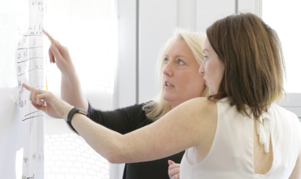 Two women at a large whiteboard point at items on the board and discuss the content