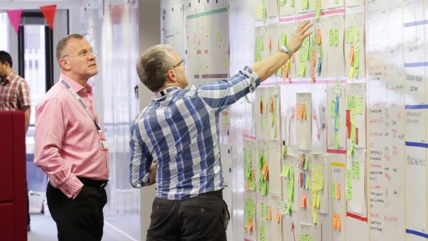 Two male colleagues talking at a whiteboard pointing at a group of Post-It Notes on the wall 