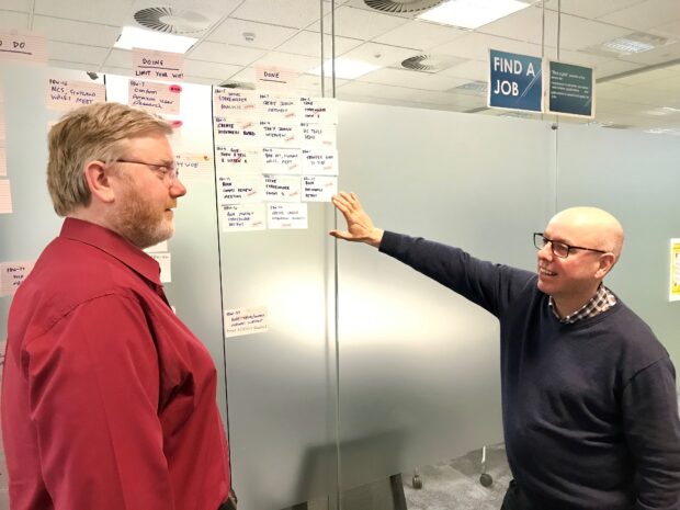 Colleagues Barry and Sean discussing the Find a Job service stood at a working board