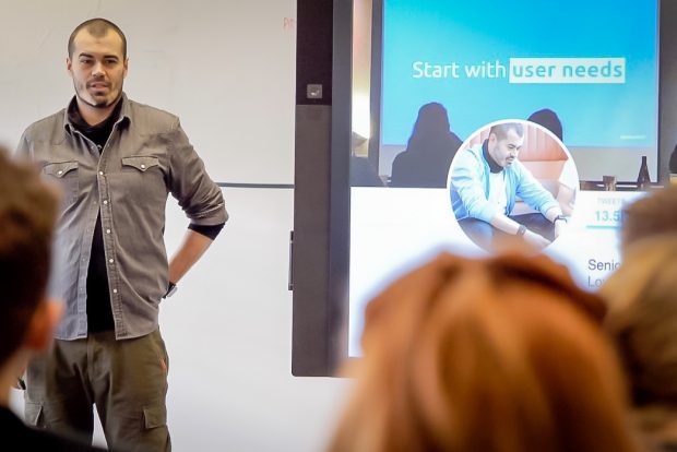 Craig Abbott, DWP Digital's Head of Accessibility stood up giving a talk on accessibility in service design in front of an audience of people, with a display screen behind him with the words 'Start with user needs' on the screen