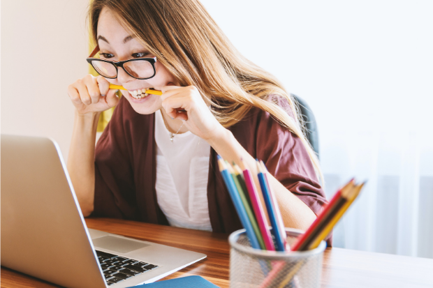 woman bites a pencil while sat at a desk in front of a laptop