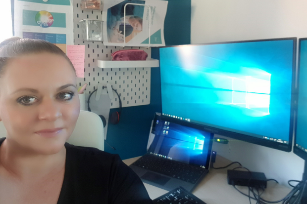 A woman sits at a desk in front of a laptop and screen