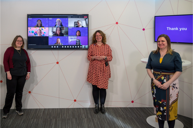 Three women stand smiling in front of a television screen that features eight women's faces