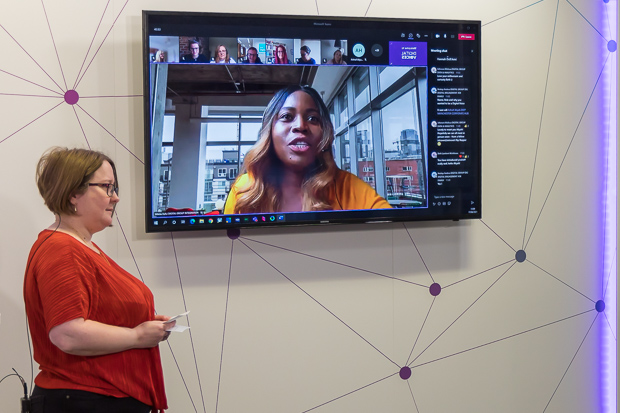 A woman in a red top stands in front of a large screen, where another woman is talking on a video call