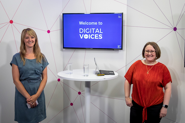 Two women stand smiling in front of a desk and a blue television screen. One is wearing a blue dress, the other has glasses and is wearing a red top and black trousers. 