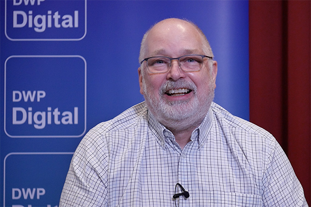 Simon sits smiling in front of a blue screen with the DWP Digital logo printed on it