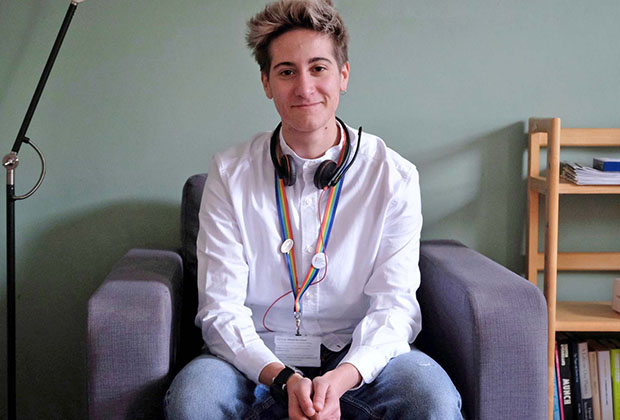 Image shows Leo say in a chair wearing a white shirt and rainbow lanyard