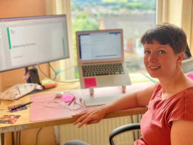 Image of Gemma Elsworth sitting at her desk looking at the camera with two screens in the background