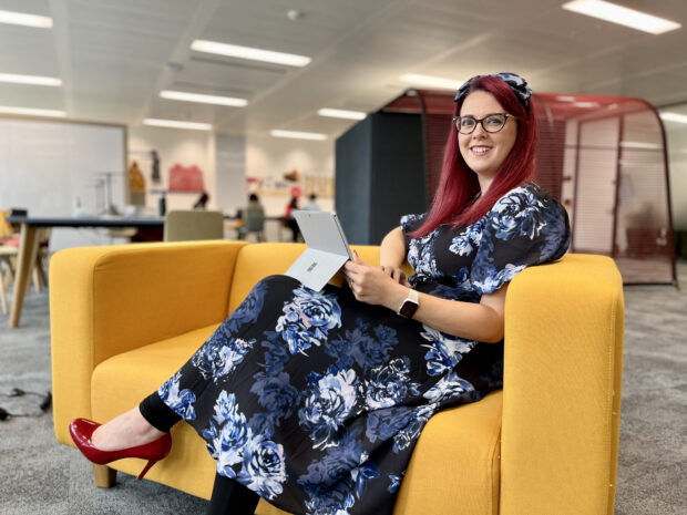 Woman sat on yellow sofa with laptop smiling to camera.