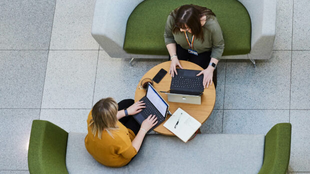 Two females working in an open air office collaborating around laptops.